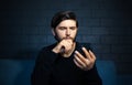 Portrait of young thoughtful man, looking in smartphone, sitting on sofa. Background of black brick wall. Royalty Free Stock Photo