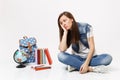 Portrait of young unhappy woman student in denim clothes resting cheek on hand sitting looking on globe backpack school