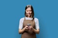 Portrait of young woman in apron holding smartphone in hands on blue background
