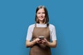 Portrait of young woman in apron holding smartphone in hands on blue background Royalty Free Stock Photo