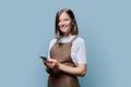 Portrait of young woman in apron holding smartphone in hands on blue background Royalty Free Stock Photo