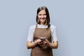 Portrait of young woman in apron holding smartphone in hands on gray background