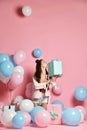 Portrait of young woman in birthday party holding gift box with gift present on pastel pink background with colorful air baloons.