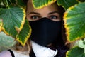 Portrait of the young woman in a black protective mask with a framed face of natural tree leaves.