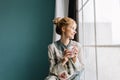 Portrait of young woman with blonde hair drinking coffee or tea next to big window, smiling, enjoying happy morning at Royalty Free Stock Photo