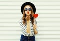 Portrait young woman blowing red lips sending sweet air kiss with red heart shaped lollipop in black round hat on white wall