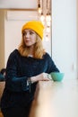 Portrait of young woman with blue eyes and blond hair in a yellow knitting hat is drinking coffee in a cafe. Royalty Free Stock Photo