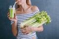 Portrait of a young woman with a bunch of celery in one hand and a glass of celery juice in the other. dark background, studio