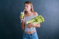 Portrait of a young woman with a bunch of celery in one hand and a glass of celery juice in the other. dark background, studio