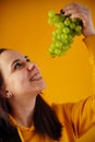 Portrait of young woman with bunch of grapes on yellow background. Close up of female opens mouth, going to eat green