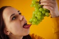 Portrait of young woman with bunch of grapes on yellow background. Close up of female opens mouth, going to eat green