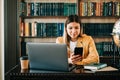 Portrait of young woman college student using mobile phone, preparing for test exam, writing essay doing homework in the library Royalty Free Stock Photo