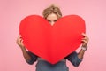 Portrait of young woman with curly hair hiding face behind big red heart and looking at camera with curious prying eyes