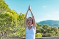 Portrait of serious young woman practicing yoga outdoors with hands raised up against blue sky and green trees