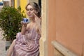 Portrait of young woman with fair hair wearing summer pink striped dress, sitting on step, enjoying aroma of coffee. Royalty Free Stock Photo