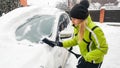 Close up portrait of young woman in green coat is getting ready to clean snow off a white car using a snow brush.