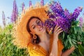 Portrait of young woman holding bouquet of lupin flowers on summer meadow. Stylish girl wearing straw hat