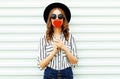 Portrait young woman kissing red heart shaped lollipop or hides her lips in black round hat, white striped shirt on white wall Royalty Free Stock Photo