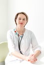 Portrait of a young woman professional doctor with short haircut, in a bright medical room, in a white coat with a phonoscope on