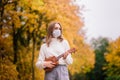 Portrait young woman in protective mask playing ukulele guitar in autumn park, healthy travel