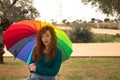 Portrait of young woman, red hair, freckles, with a rainbow umbrella, happy, in an outdoor park. Concept color, happiness, well-