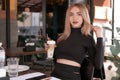 Portrait of young woman with red lips wears black clothes and holds cup of cappuccino. Young attactive girl in cafe