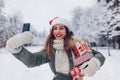 Portrait of young woman in Santa hat taking selfie on smartphone in snowy winter park holding heap of Christmas presents