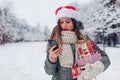 Portrait of young woman in Santa hat using phone on smartphone in snowy winter park holding heap of Christmas presents Royalty Free Stock Photo