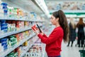 Portrait of young woman scans the QR code on a package of yogurt. In the background, a supermarket with visitors in a Royalty Free Stock Photo