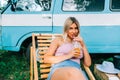Portrait of young woman sitting on wooden chair near van and drinking lemonade outdoors in nature. Enjoying summer, travel concept Royalty Free Stock Photo