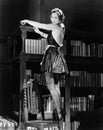 Portrait of a young woman standing on a chair and dusting a bookshelf in a outfit