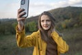 Portrait of Young woman taking selfie on smartphone, mountain background. Girl in yellow jacket enjoys of autumn nature