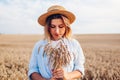 Portrait of young woman walking among wheat in summer field wearing straw hat holding bundle of ripe wheat