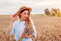 Portrait of young woman walking among wheat in summer field wearing straw hat holding bundle of ripe wheat