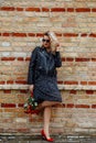 Portrait of young woman wearing black dress, leather jacket, holding bouquet of red roses, leaning against brick wall. Royalty Free Stock Photo