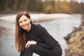 Portrait of young woman wearing black jeans and top, leaning, her long hair falling down, smiling. Blurred river in background Royalty Free Stock Photo