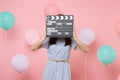 Portrait of young woman wearing blue dress covering face with classic black film making clapperboard on pink background