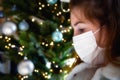 Portrait of a young woman wearing protective face mask and looking sad for Covid-19 with Christmas tree
