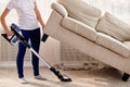 Portrait of young woman in white shirt and jeans cleaning carpet under sofa with vacuum cleaner in living room, copy space. Royalty Free Stock Photo