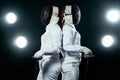 Young fencer athlete wearing mask and white fencing costume. holding the sword on black background with lights.