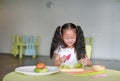 Portraits of happy Asian child girl slicing cucumber vegetable on chopping board at play room. Kid play chef cooking