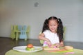 Portraits of happy Asian child girl slicing cucumber vegetable on chopping board at play room. Kid play chef cooking