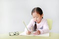 Portraits of little Asian child girl write in a book or notebook with pencil sitting on kid chair and table against white Royalty Free Stock Photo