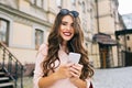 Portraut of cute girl with long curly hair and phone in hands smiling to camera in city on building background