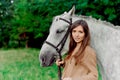 A portret of a young woman, a jockey-girl, that leads a saddled grey horse on grass.