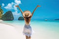 Portriat of young beautiful woman raised hands and wearing a hat and white dress staying on a tropical beach near sea