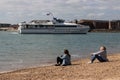 Two women sitting together on a pebble beach in the summer with a ferry passing Royalty Free Stock Photo