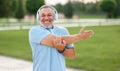Positive confident senior sportsman wearing wireless headphones doing arm stretching in park outside