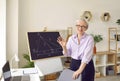 Positive adult gray haired woman teacher stands next to table with laptop and blackboard Royalty Free Stock Photo