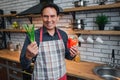 Positive adult man stand alone in kitchen. He smiles to camera. Guy hold orange pepper and green onion. Royalty Free Stock Photo